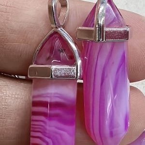 May include: Two pink and white agate crystal pendants with silver-colored metal caps and loops. The crystals have a layered, striped appearance. The pendants are displayed against a neutral background.
