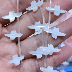 Może przedstawiać: Szereg białych pereł z masy perłowej w kształcie ptaków na przezroczystym sznurku elastycznym. Perły są nawleczone w powtarzający się wzór.