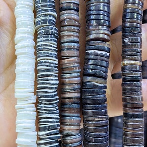 以下が含まれることがあります： 異なるパターンを持つ貝殻ビーズの4つのストランド。ビーズは積み重ねられ、垂直に配置されています。ストランドは白、黒と白のストライプ、茶色と白のストライプ、濃い茶色です。