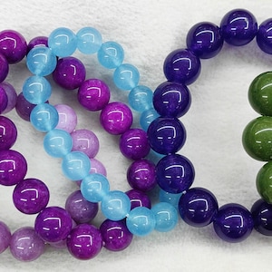 Può includere: Una collezione di braccialetti di perline in vari colori, tra cui rosa, viola, blu, verde e giallo. Ogni braccialetto è composto da perline rotonde, creando un'esposizione colorata e accattivante. I braccialetti sono disposti uno accanto all'altro.