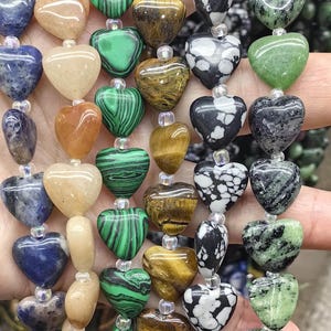 May include: A selection of heart-shaped gemstone beads in a variety of colours and patterns. The beads include shades of green, brown, black, white, blue, and tan. The gemstones are strung together with clear string, creating a unique jewellery design.