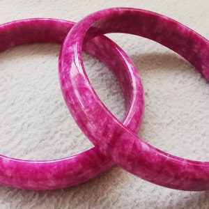 Puede incluir: Dos brazaletes de jade rosa con un patrón de mármol blanco. Los brazaletes están apilados uno encima del otro.