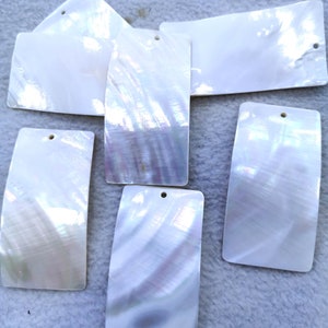 May include: Six white iridescent mother of pearl shell blanks with a hole drilled in the top. The blanks are rectangular and oval shaped.