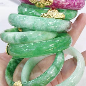 Könnte beinhalten: Ein Stapel Jade-Armreifen in verschiedenen Grüntönen und ein rosa Armreif. Jeder Armreif hat einen goldfarbenen floralen Akzent mit einem kleinen Edelstein. Die Armreifen sind glatt und poliert und zeigen die natürlichen Muster des Jades.
