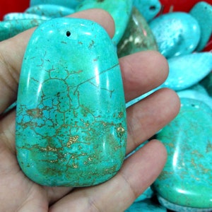 May include: A smooth, oval-shaped turquoise stone with a hole drilled through the top for stringing. The stone has a bright blue-green colour with brown veins and markings.