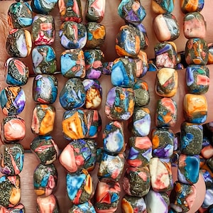 May include: A strand of colorful, multi-faceted gemstone beads. The beads are square-shaped and have a variety of colors, including blue, green, orange, and purple. The beads are strung on a clear string.