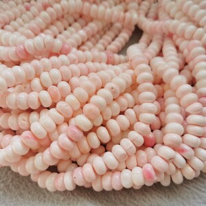 Peut inclure: Un brin de perles de corail rose pâle. Les perles sont petites et rondes, avec une forme légèrement irrégulière. Elles sont enfilées sur un fil transparent.