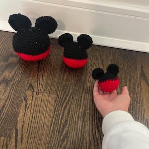 Op de afbeelding: Drie gehaakte Mickey Mouse-hoofden in verschillende maten. De hoofden zijn zwart met rode bodems en zwarte oren. Het grootste hoofd staat links, het middelste hoofd in het midden en het kleinste hoofd wordt rechts door een hand vastgehouden.
