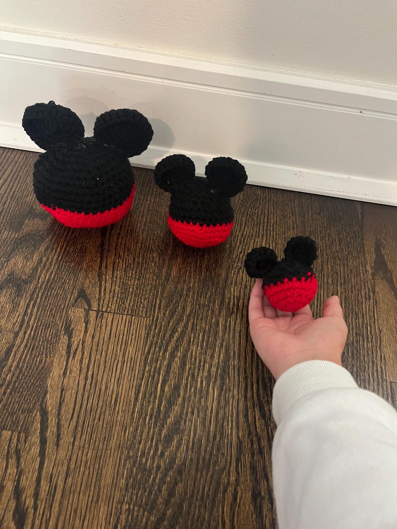 May include: Three crocheted Mickey Mouse heads in varying sizes. The heads are black with red bottoms and black ears. The largest head is on the left, the medium head is in the middle, and the smallest head is being held by a person's hand on the right.