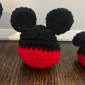 May include: Crocheted Mickey Mouse head with black ears and a red and black face. The handmade item is likely a toy or decorative piece, showcasing a classic cartoon character design. The ears are round and prominent.