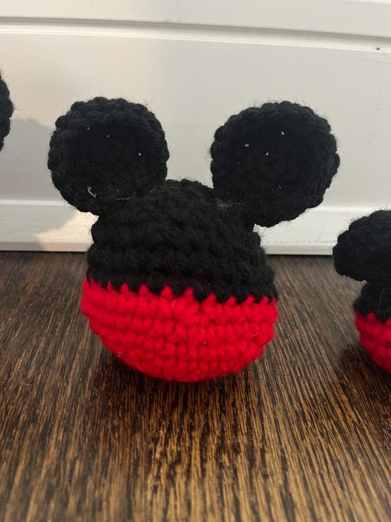 May include: Crocheted Mickey Mouse head with black ears and a red and black face. The handmade item is likely a toy or decorative piece, showcasing a classic cartoon character design. The ears are round and prominent.