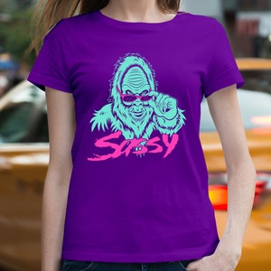 May include: Purple t-shirt with a blue and pink graphic of a sasquatch wearing sunglasses and the word "Sassy" in pink.