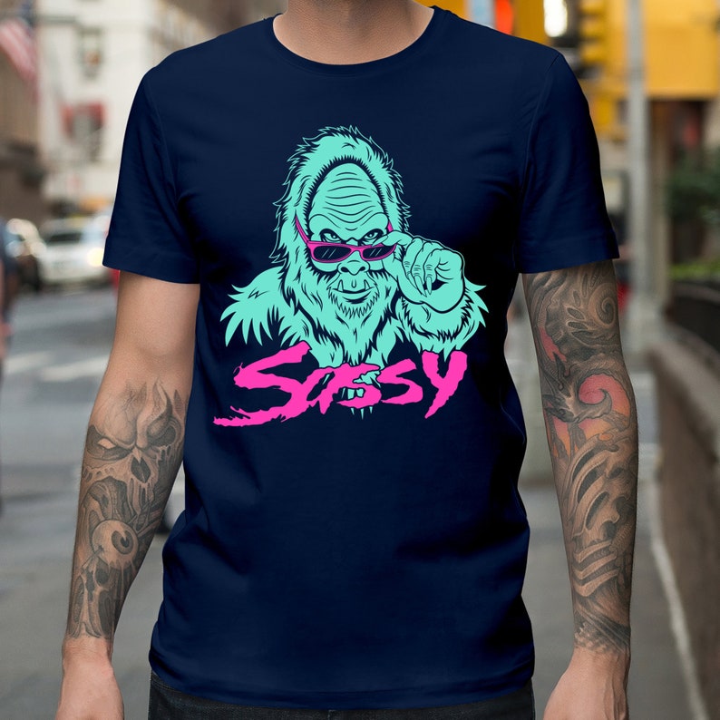 May include: A navy blue t-shirt with a graphic of a blue sasquatch wearing pink sunglasses and the word "Sassy" in pink and blue lettering.