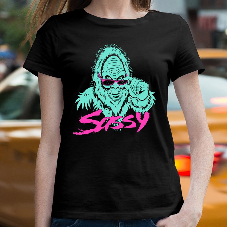 May include: A black t-shirt with a turquoise blue illustration of a sasquatch wearing pink sunglasses and the word "Sassy" in pink letters.
