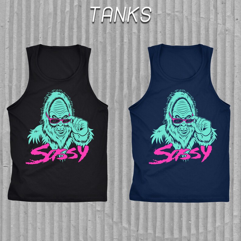 May include: Two sleeveless shirts with a graphic print of a blue sasquatch wearing pink sunglasses and the word "Sassy" in pink. The shirt on the left is black and the shirt on the right is navy blue.