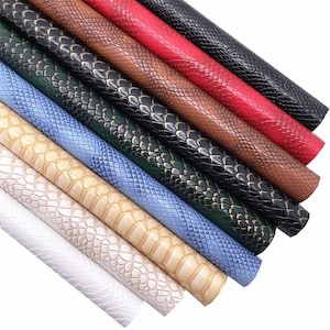 Op de afbeelding: Een verzameling kunstleren vellen in verschillende kleuren, waaronder rood, zwart, bruin en blauw. Elk vel heeft een reptielenhuidtextuur, waardoor een realistisch reliëfpatroon ontstaat. De vellen zijn opgerold en tonen de verschillende kleuren en texturen.