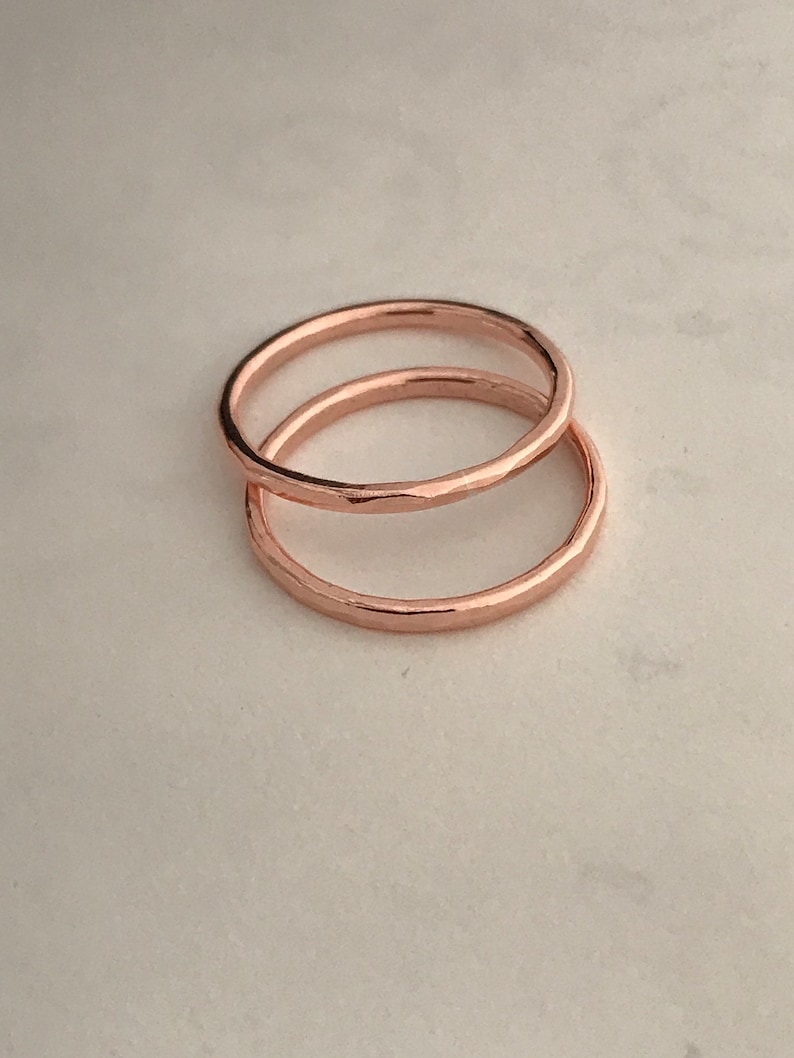 May include: Two thin rose gold rings stacked on top of each other. The rings are smooth and have a hammered texture.