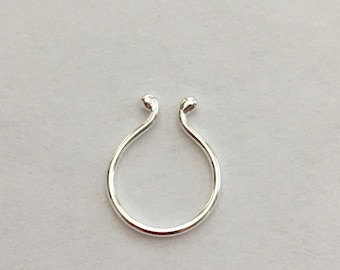 Puede incluir: Un anillo de nariz plateado con un diseño en forma de U. Los extremos del anillo se curvan hacia arriba y tienen pequeñas puntas redondeadas. La joya está sobre un fondo liso y claro.