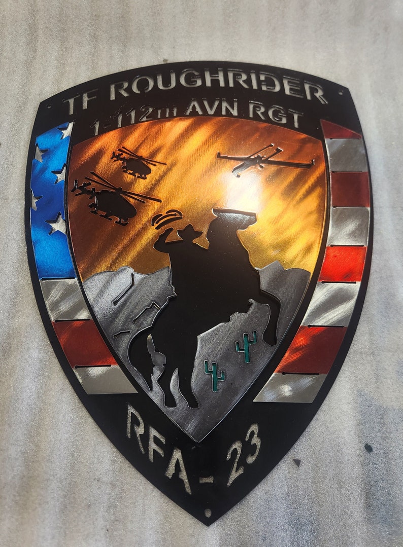 K&ouml;nnte beinhalten: Ein Metallschild mit einem amerikanischen Flaggen-Design. Das Schild zeigt eine Silhouette eines Cowboys, der ein Pferd reitet, mit Kakteen im Hintergrund. Der Text "TF ROUGH RIDER 1-112TH AVN RGT RFA-23" ist auf dem Schild zu sehen.