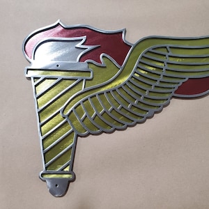 Puede incluir: Una decoración de pared de metal dorado y plateado con un diseño estilizado de ala y antorcha. La antorcha está rayada de oro y tiene una llama roja.