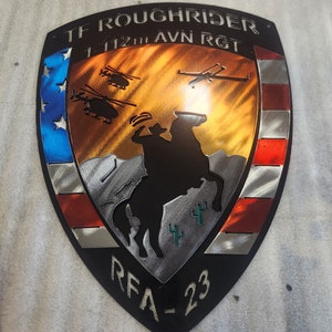 K&ouml;nnte beinhalten: Ein schwarzer Metallschild mit einem rot-wei&szlig;-blauen amerikanischen Flaggen-Design. Das Schild zeigt eine Silhouette eines Cowboys, der ein Pferd reitet, mit Kakteen im Hintergrund. Der Text "TF ROUGH RIDER 1-112TH AVN RGT RFA-23" ist auf dem Schild abgebildet.