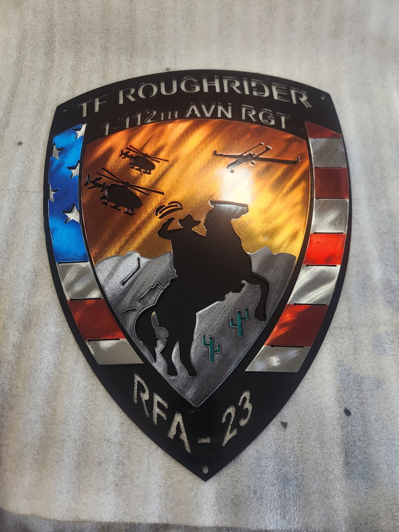 K&ouml;nnte beinhalten: Ein schwarzer Metallschild mit einem rot-wei&szlig;-blauen amerikanischen Flaggen-Design. Das Schild zeigt eine Silhouette eines Cowboys, der ein Pferd reitet, mit Kakteen im Hintergrund. Der Text "TF ROUGH RIDER 1-112TH AVN RGT RFA-23" ist auf dem Schild abgebildet.