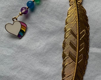 May include: A gold feather bookmark with a rainbow heart charm. The charm has a white heart with a rainbow stripe design. The bookmark is attached to a chain with colorful beads.