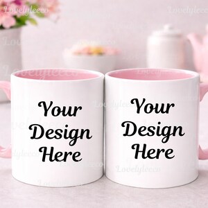 May include: Two white ceramic mugs with pink handles and interiors. Each mug has the text "Your Design Here" in black. The mugs are displayed on a light-colored surface, with a blurred background of pink and white objects.