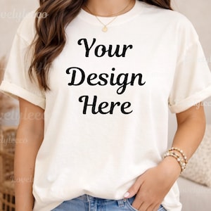 May include: Cream-colored t-shirt with rolled-up sleeves. The text "Your Design Here" is printed in black script across the front. The model is wearing a gold necklace and bracelets.
