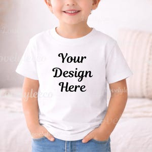 May include: A white t-shirt with the text "Your Design Here" in black script. The shirt is being worn by a child, who is wearing blue jeans. The child has their hands in their pockets.
