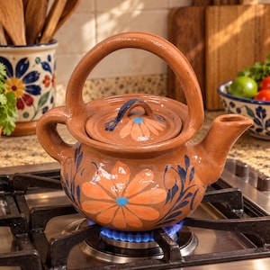 May include: A brown clay teapot with a high arched handle and a matching lid. The teapot features hand-painted orange flower motifs with blue centers and dark blue leafy accents on the body and lid. It sits on a metal gas stove burner.