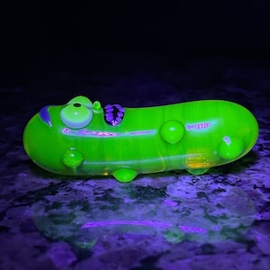 May include: A bright green glass pickle with a face and a purple swirl design. The pickle is lying on a dark surface.