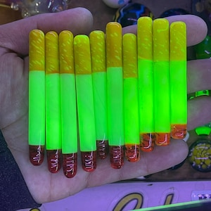 May include: A collection of neon green and yellow glass smoking accessories. Each piece has a yellow top, a neon green body, and a red base. The items are held in a hand, with other glass art pieces in the background.