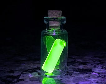 May include: A small, clear glass bottle with a cork stopper, containing a glowing, neon green cylindrical object. The bottle is reflected on a dark, textured surface, creating a vibrant, otherworldly effect. The background is black.