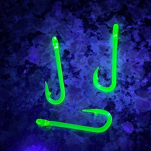 Puede incluir: Tres anzuelos de pesca verdes brillan intensamente bajo una luz negra. Los anzuelos están hechos de un material translúcido y tienen una punta afilada.