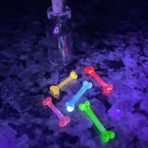 Puede incluir: Seis encantos de plástico en forma de hueso que brillan en la oscuridad, en varios colores, incluidos amarillo, verde, naranja y rojo, están esparcidos alrededor de una pequeña botella de vidrio con un tapón de corcho.