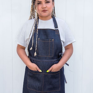 May include: A person wearing a white t-shirt and a dark blue denim apron with a large pocket and adjustable straps. The apron has a small red label with white text.