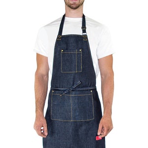 May include: A dark blue denim apron with a large pocket and adjustable straps. The apron has yellow stitching and brass hardware.