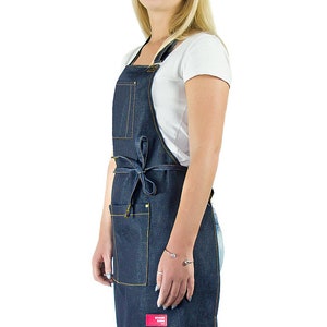 May include: A denim apron with yellow stitching and a red logo that says "Brand Name". The apron has a large pocket on the front and ties at the waist.