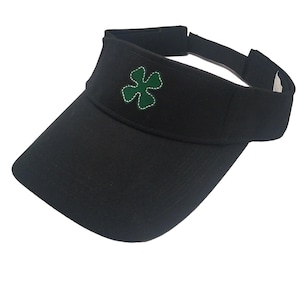 May include: Black visor featuring a green four-leaf clover emblem with a white outline. The visor has an adjustable strap and a curved peak. A practical accessory for shielding the eyes from the sun.