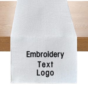 Puede incluir: Camino de mesa rectangular blanco con las palabras "Embroidery Text Logo" en negro. El camino de mesa está hecho de una tela texturizada y se muestra sobre una mesa de madera.