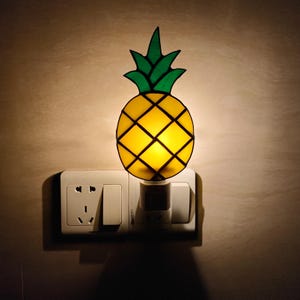 May include: A decorative night light shaped like a pineapple. The pineapple has a yellow body with a black grid pattern and green leaves. It is plugged into a white electrical outlet.