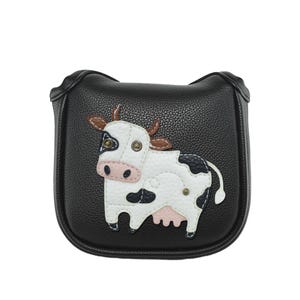 May include: Black golf club head cover with a cow design. The cover has a black background with a white cow with black spots, brown horns, and pink accents. The cover is made of a textured material.