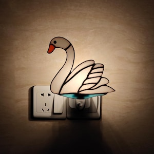 May include: A swan-shaped plug-in night light featuring white stained glass panels with black outlines, a bright orange beak, and a small teal accent at the base. The light is plugged into a white wall outlet, emitting a soft, warm glow from within.