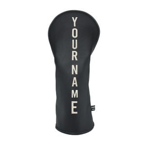 May include: Black golf club head cover with the text "YOUR NAME" in white capital letters. The cover is made of a leather-like material and has a rounded top. A small tag is visible on the side.