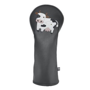 Puede incluir: Una funda negra para la cabeza del palo de golf con un diseño de vaca. La vaca es blanca con manchas negras, una ubre rosa y orejas marrones. La funda está hecha de un material texturizado.