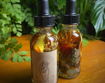 May include: Two small glass bottles filled with herbs, crystals, and liquid. The bottles have black dropper tops and brown labels that say "Abundance Oil" with a hand holding a gemstone.
