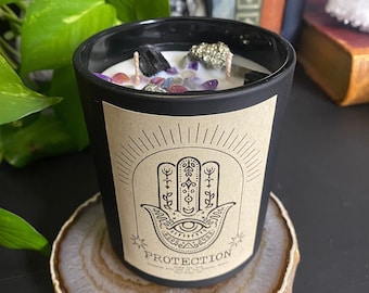 May include: Black candle with a brown label featuring a hamsa hand symbol and the word "PROTECTION". The candle is filled with crystals and has a white wax top.