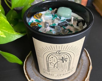May include: Black candle with a brown label that says "Road Opener" and a design of a moon, stars, and a palm tree in an archway. The candle is filled with various crystals and is sitting on a brown agate slice.