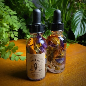 May include: Two clear glass bottles with dropper tops filled with dried flowers, herbs, and crystals. The bottles have brown paper labels with the text "ENERGETIC BALANCING OIL" and a drawing of a woman with a scale.